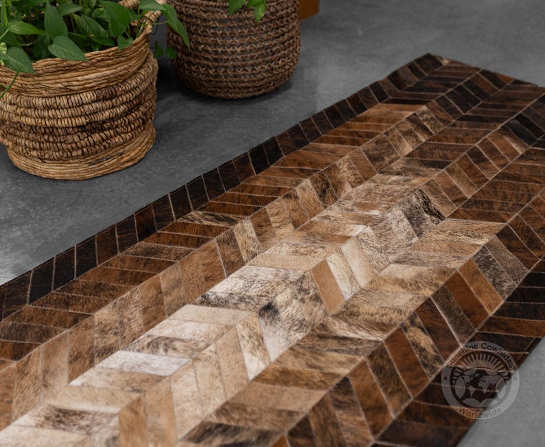 May include: A rectangular rug with a chevron pattern, featuring various shades of brown and tan. The rug is made of a natural material, possibly cowhide, and has a textured appearance. The design creates a visual flow across the surface.