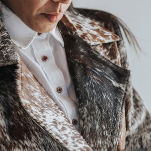 May include: A cowhide jacket with a white collared shirt underneath. The jacket features a mix of brown, black, and white patterns. The shirt has brown buttons. The jacket is zipped up.
