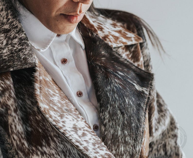 May include: A cowhide jacket with a white collared shirt underneath. The jacket features a mix of brown, black, and white patterns. The shirt has brown buttons. The jacket is zipped up.