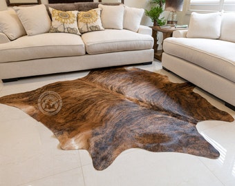 May include: A brown and black cowhide rug with a natural, irregular shape. The rug is laid out on a light beige tiled floor in front of two white sofas.