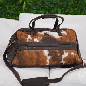 May include: A duffel bag crafted from brown and white cowhide with dark brown leather trim. The bag features two top handles, a detachable shoulder strap with a padded section, and a horizontal zippered pocket across the front center panel.