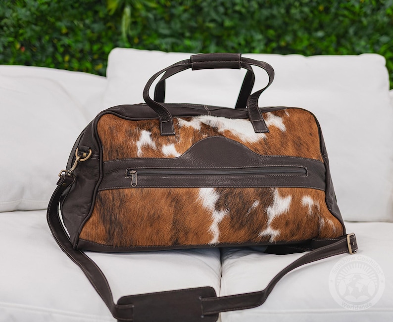 May include: A duffel bag crafted from brown and white cowhide with dark brown leather trim. The bag features two top handles, a detachable shoulder strap with a padded section, and a horizontal zippered pocket across the front center panel.