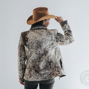 May include: A brown cowboy hat and a jacket with a cowhide pattern. The jacket has a mix of brown, white, and black colors. The person is wearing dark jeans. The image is taken from the back.