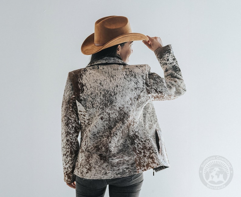 May include: A brown cowboy hat and a jacket with a cowhide pattern. The jacket has a mix of brown, white, and black colors. The person is wearing dark jeans. The image is taken from the back.