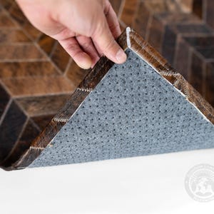 May include: A brown and black cowhide rug with a geometric pattern. The rug's underside is gray with small holes. The edge of the rug is being lifted to show the underside. The rug is on a white surface.