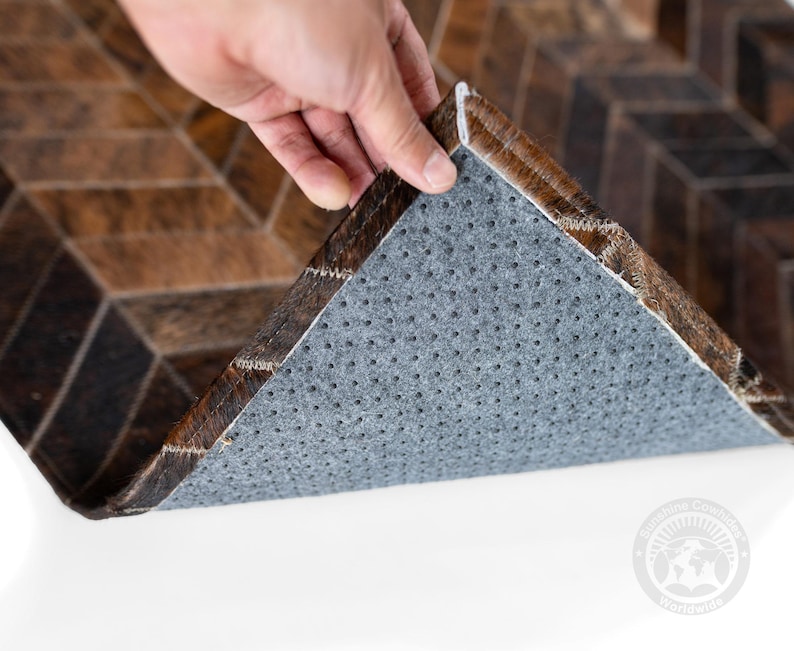 May include: A brown and black cowhide rug with a geometric pattern. The rug's underside is gray with small holes. The edge of the rug is being lifted to show the underside. The rug is on a white surface.