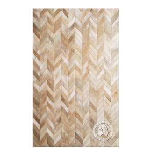 Może przedstawiać: Prostokątny dywan z wzorem w jodełkę. Dywan wykonany jest ze skóry krowiej w naturalnych odcieniach, z odcieniami beżu, jasnobeżowego i jasnobrązowego. Wzór to powtarzająca się seria jodełek, tworząca teksturowaną powierzchnię. Dywan ma logo w prawym dolnym rogu.