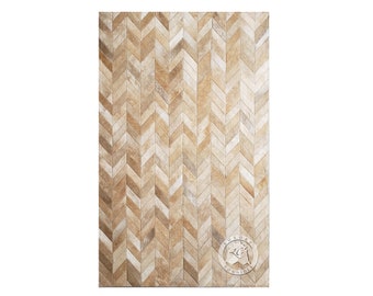 May include: A rectangular rug with a chevron pattern. The rug is made of natural-toned cowhide, with shades of beige, tan, and light brown. The pattern is a repeating series of chevrons, creating a textured surface. The rug has a logo in the bottom right corner.