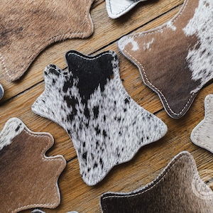 May include: A selection of cowhide coasters in diverse patterns and colours, including brown, black, and white. Each coaster has a distinctive shape and is stitched with white thread. The coasters are arranged on a wooden surface.