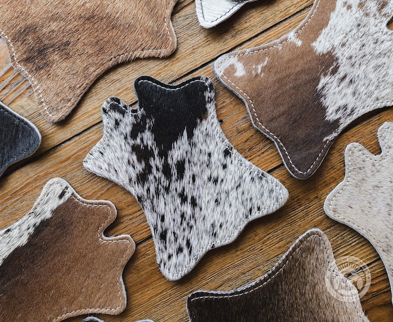 May include: A selection of cowhide coasters in diverse patterns and colours, including brown, black, and white. Each coaster has a distinctive shape and is stitched with white thread. The coasters are arranged on a wooden surface.