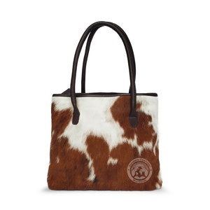 May include: A brown and white cowhide tote bag with brown leather handles. The bag has a brown leather trim and a small brown leather tag with the text "Brisbane Cowhide Worldwide".