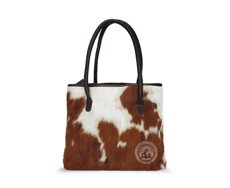 May include: A brown and white cowhide tote bag with brown leather handles. The bag has a brown leather trim and a small brown leather tag with the text "Brisbane Cowhide Worldwide".