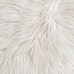 May include: A close-up view of long, soft, off-white faux fur material. The texture consists of dense, flowing synthetic fibers that create a shaggy, layered appearance with subtle variations in tone, ranging from cream to bright white throughout.