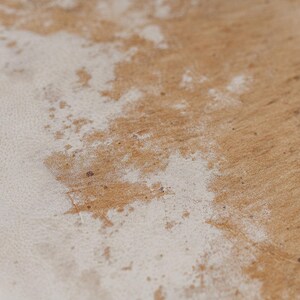 May include: A close-up view of a textured surface featuring a natural light brown wood grain pattern partially covered by an irregular, weathered layer of white paint or finish that reveals the underlying wood texture in various spots.