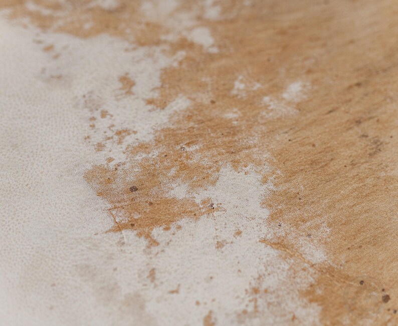 May include: A close-up view of a textured surface featuring a natural light brown wood grain pattern partially covered by an irregular, weathered layer of white paint or finish that reveals the underlying wood texture in various spots.