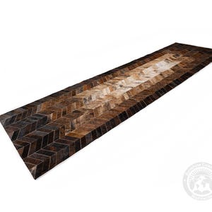 May include: A long, rectangular rug with a chevron pattern. The rug features a gradient of brown tones, from dark chocolate to light tan, creating a visually appealing design. The rug is made of a natural material, likely cowhide, and is suitable for a hallway or entryway.