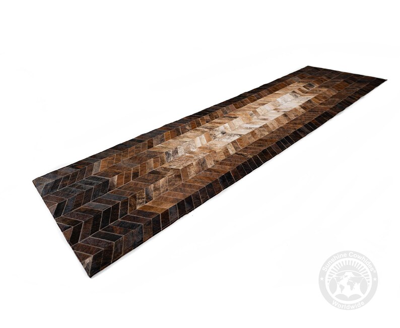 May include: A long, rectangular rug with a chevron pattern. The rug features a gradient of brown tones, from dark chocolate to light tan, creating a visually appealing design. The rug is made of a natural material, likely cowhide, and is suitable for a hallway or entryway.