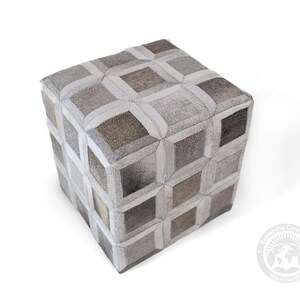 May include: A cube-shaped ottoman with a geometric pattern. The ottoman is upholstered in a patchwork of gray, brown, and white cowhide pieces. The design features a repeating pattern of squares and rectangles, creating a textured surface.