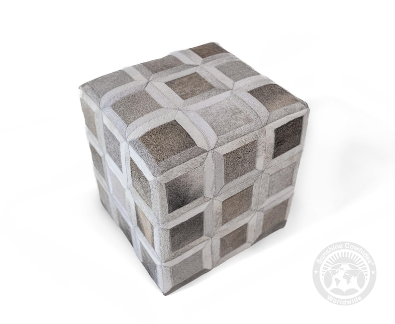 May include: A cube-shaped ottoman with a geometric pattern. The ottoman is upholstered in a patchwork of gray, brown, and white cowhide pieces. The design features a repeating pattern of squares and rectangles, creating a textured surface.