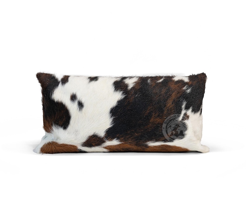 May include: A rectangular cowhide pillow in brown, black, and white. The pillow features a natural cowhide pattern with variations in color and texture. The pillow is shown against a white background. A small, circular logo is visible on the pillow.