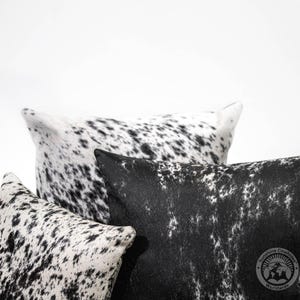 May include: Three black and white cowhide pillows with a black and white spotted pattern. The pillows are arranged in a stack with the largest pillow on the bottom and the smallest pillow on top. The pillows are made of soft, luxurious cowhide and are perfect for adding a touch of rustic elegance to any room.