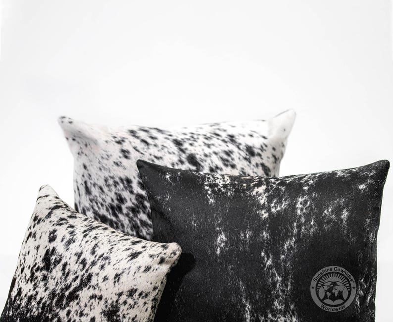 May include: Three black and white cowhide pillows with a black and white spotted pattern. The pillows are arranged in a stack with the largest pillow on the bottom and the smallest pillow on top. The pillows are made of soft, luxurious cowhide and are perfect for adding a touch of rustic elegance to any room.
