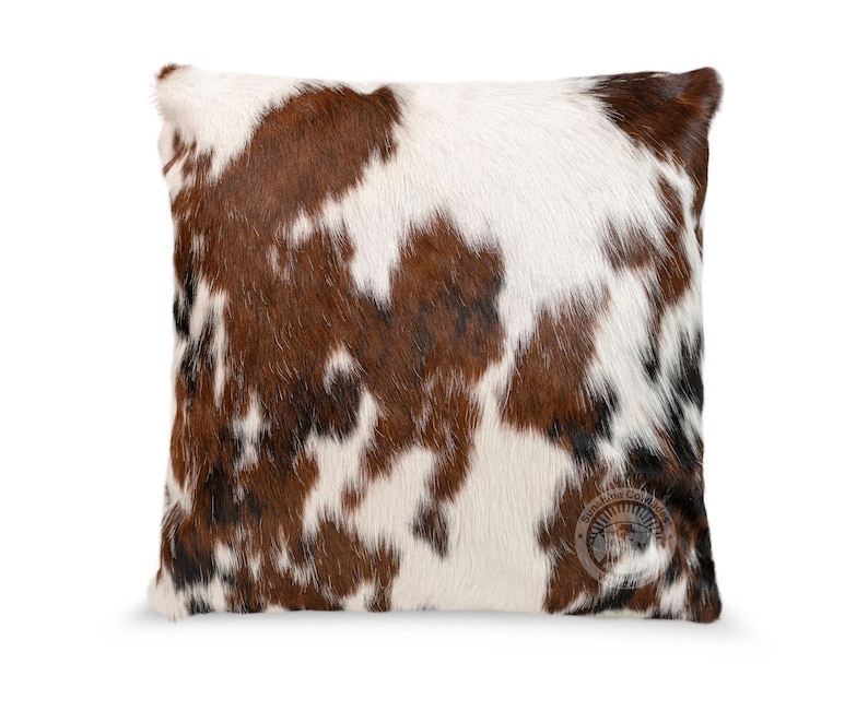 May include: A square throw pillow featuring a natural cowhide pattern with patches of white, dark brown, and black fur. The surface has a textured, hair-on-hide appearance. A circular logo with the text Sunshine Cowhides is visible in the bottom right.