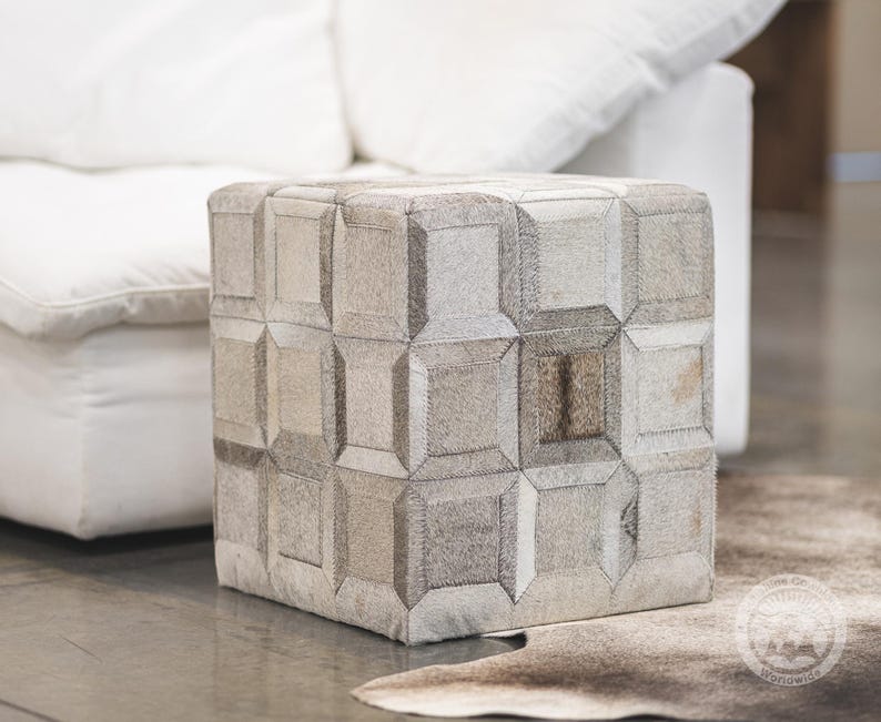 May include: A square ottoman with a geometric patchwork design. The ottoman is made of neutral-toned, textured materials, with a pattern of squares and rectangles. It sits on a light-colored floor, with a white sofa in the background.