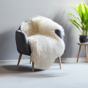 Puede incluir: Un sill&oacute;n gris con patas de madera est&aacute; cubierto con una alfombra de piel de oveja suave y blanquecina. Una planta en maceta con hojas verdes grandes se encuentra sobre una peque&ntilde;a mesa auxiliar de madera.