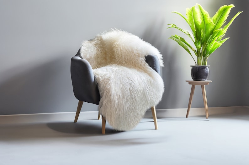 Puede incluir: Un sill&oacute;n gris con patas de madera est&aacute; cubierto con una alfombra de piel de oveja suave y blanquecina. Una planta en maceta con hojas verdes grandes se encuentra sobre una peque&ntilde;a mesa auxiliar de madera.