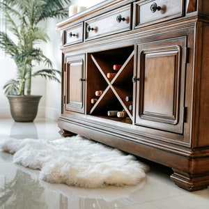 Puede incluir: Un mueble bar de madera marr&oacute;n oscuro con botellero incorporado, cajones y puertas de armario. El mueble est&aacute; sobre un suelo blanco brillante, con una alfombra de piel sint&eacute;tica blanca delante. Una planta en maceta est&aacute; al fondo.
