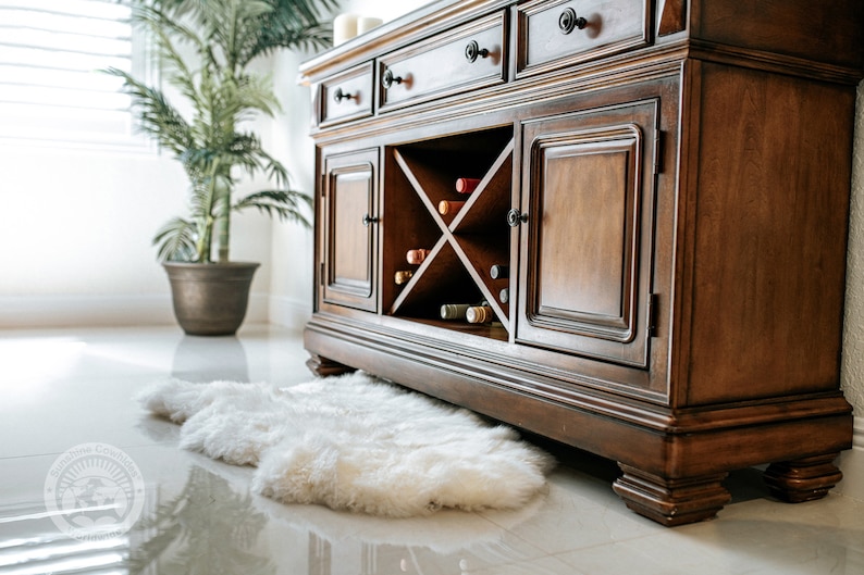Puede incluir: Un mueble bar de madera marr&oacute;n oscuro con botellero incorporado, cajones y puertas de armario. El mueble est&aacute; sobre un suelo blanco brillante, con una alfombra de piel sint&eacute;tica blanca delante. Una planta en maceta est&aacute; al fondo.