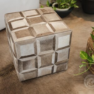 Pu&ograve; includere: Un pouf quadrato con un motivo geometrico in pelle marrone chiaro e bianco. Il pouf &egrave; su un tappeto marrone chiaro.