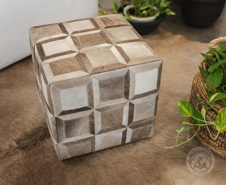 Pu&ograve; includere: Un pouf quadrato con un motivo geometrico in pelle marrone chiaro e bianco. Il pouf &egrave; su un tappeto marrone chiaro.