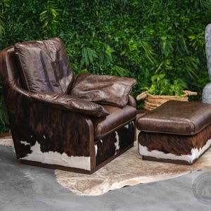 May include: A brown leather armchair with a white and brown cowhide pattern. The armchair has a matching ottoman with a brown leather top and a white and brown cowhide base. The armchair and ottoman are sitting on a white rug.