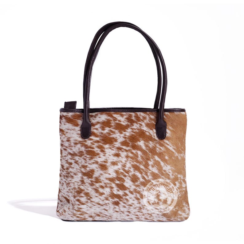 May include: A brown and white cowhide tote bag with brown leather handles and a brown leather trim. The bag has a logo on the front that says "United We Cowhide".