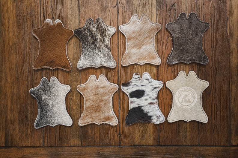 May include: Eight cowhide coasters in various colours and patterns, including brown, grey, and black, arranged on a wooden surface. Each coaster has a unique natural hide pattern and a stitched border.