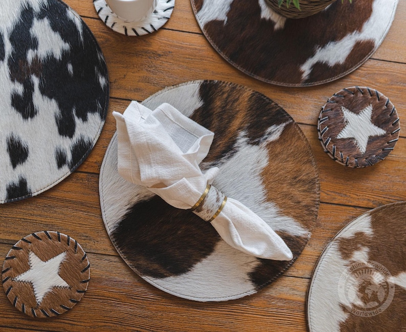 May include: A collection of circular placemats and coasters featuring brown, white, and black cowhide patterns. A white linen napkin is folded and secured with a metallic napkin ring, resting on a large round placemat with a brown and white hide design.