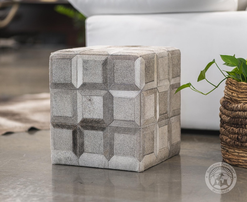 May include: A square gray and white cowhide ottoman with a geometric pattern. The ottoman is sitting on a gray floor.  The ottoman is made of genuine cowhide and is a great addition to any home.
