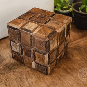 Pu&ograve; includere: Un pouf quadrato con un motivo geometrico in pelle marrone, beige e bianco. Il pouf &egrave; appoggiato su un tappeto marrone.