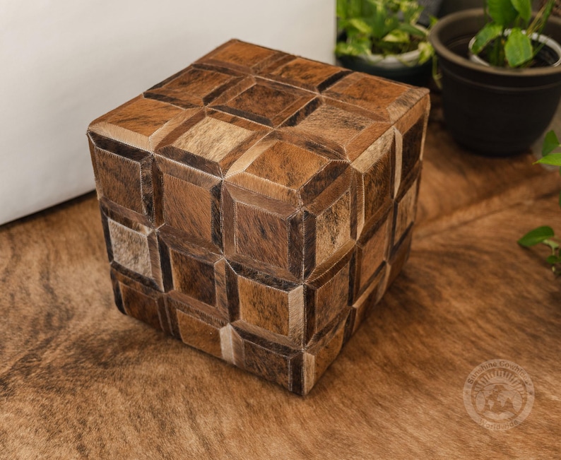 Pu&ograve; includere: Un pouf quadrato con un motivo geometrico in pelle marrone, beige e bianco. Il pouf &egrave; appoggiato su un tappeto marrone.