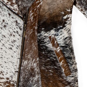 May include: A close-up of a cowhide jacket with a zipper closure. The jacket features a mix of brown, white, and black spotted patterns. The jacket has a pocket on the front and a long sleeve.