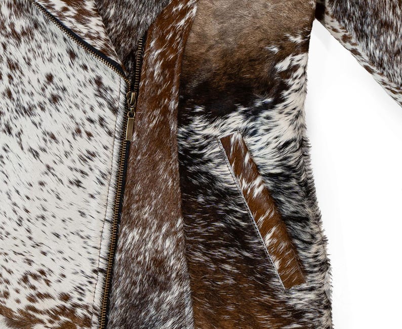 May include: A close-up of a cowhide jacket with a zipper closure. The jacket features a mix of brown, white, and black spotted patterns. The jacket has a pocket on the front and a long sleeve.