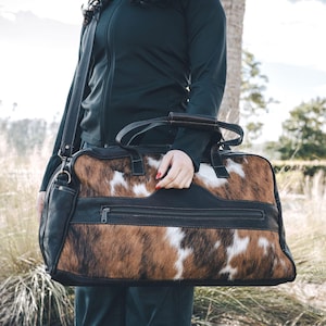 May include: A rectangular duffel bag featuring a brown, black, and white cowhide pattern on the front and back panels. The bag includes black leather trim, black leather handles, a detachable shoulder strap, and a horizontal zippered pocket.