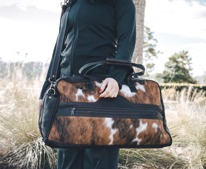 May include: A rectangular duffel bag featuring a brown, black, and white cowhide pattern on the front and back panels. The bag includes black leather trim, black leather handles, a detachable shoulder strap, and a horizontal zippered pocket.