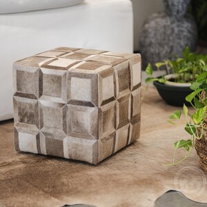Pu&ograve; includere: Un pouf quadrato con un motivo geometrico in pelle marrone chiaro e scuro. Il pouf &egrave; appoggiato su un tappeto marrone chiaro.
