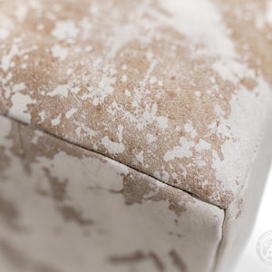 Puede incluir: Un objeto rectangular que presenta una superficie texturizada con un patr&oacute;n moteado de colores beige claro y blanco roto. El material muestra una apariencia desgastada y envejecida con parches de color irregulares distribuidos por la parte superior plana y el lateral.