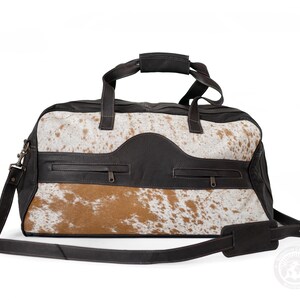 May include: A duffel bag featuring a combination of dark brown leather and white and tan cowhide fur. The bag includes two top handles, a detachable shoulder strap, and two horizontal zippered pockets on the front panel with dark metal hardware.