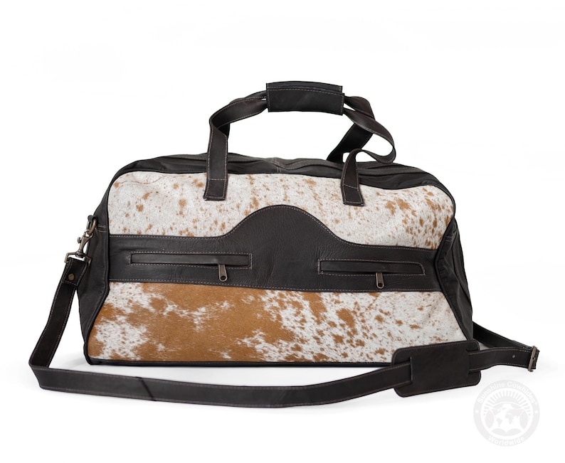 May include: A duffel bag featuring a combination of dark brown leather and white and tan cowhide fur. The bag includes two top handles, a detachable shoulder strap, and two horizontal zippered pockets on the front panel with dark metal hardware.