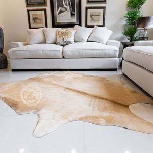 May include: A light brown cowhide rug with a natural, irregular shape. The rug is laid out on a white tiled floor in front of a white sofa and a white armchair.